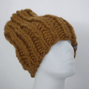 May include: A mustard-colored, ribbed knit beanie hat. The hat is made of thick yarn, with a prominent ribbed texture running horizontally across the crown. A small, gray tag is visible near the brim. The hat is displayed on a white mannequin head.