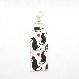 May include: A white fabric keychain with a silver clip. The fabric has a repeating pattern of black cats with red hearts.