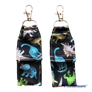 K&ouml;nnte beinhalten: Zwei schwarze Stoffschl&uuml;sselanh&auml;nger mit silbernen Clips. Der Stoff ist mit einem Muster aus bunten Dinosauriern auf schwarzem Hintergrund bedruckt. Die Schl&uuml;sselanh&auml;nger haben eine kleine Tasche auf der Vorderseite.