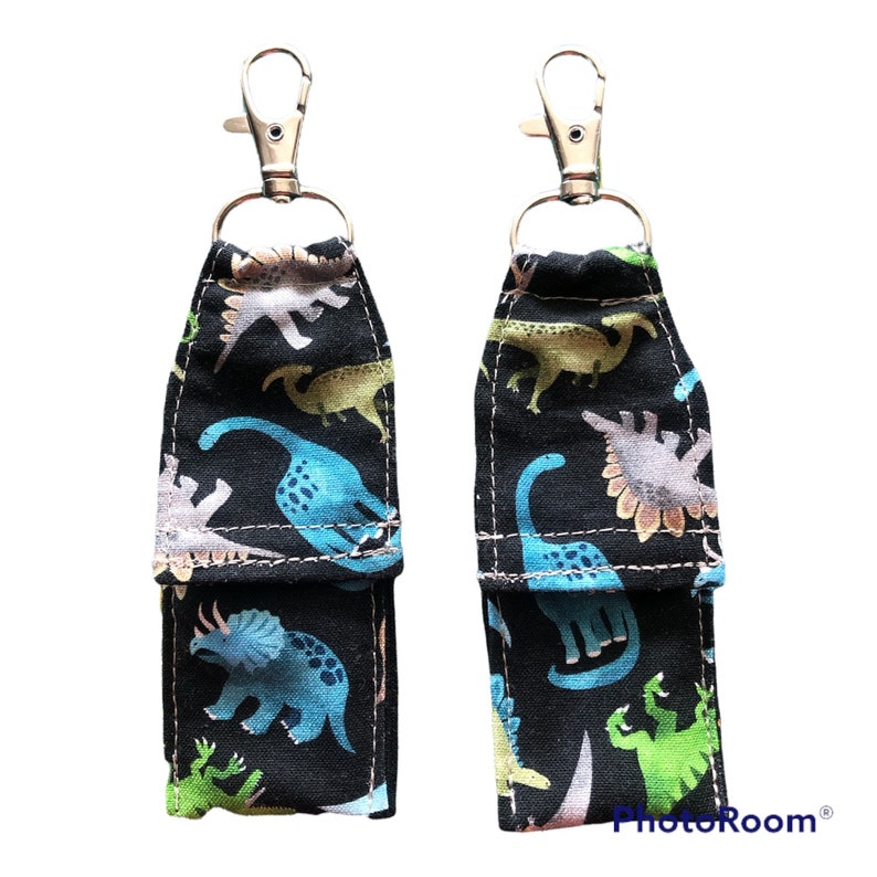 K&ouml;nnte beinhalten: Zwei schwarze Stoffschl&uuml;sselanh&auml;nger mit silbernen Clips. Der Stoff ist mit einem Muster aus bunten Dinosauriern auf schwarzem Hintergrund bedruckt. Die Schl&uuml;sselanh&auml;nger haben eine kleine Tasche auf der Vorderseite.