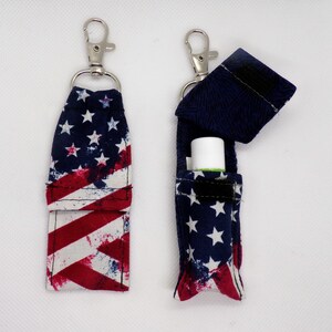 May include: Two fabric lip balm holders featuring a red, white, and blue American flag print. Each holder has a silver metal swivel lobster clasp attached to a ring. One holder is closed, while the other is open, revealing a white lip balm tube.