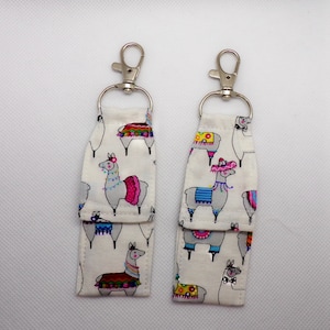 May include: Two fabric keychains with silver-colored clasps. The keychains are white with a repeating pattern of gray llamas wearing colorful accessories. The llamas have pink, blue, and yellow accents. The keychains are rectangular with a folded bottom.