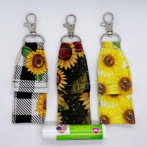 May include: Three keychains with fabric covers featuring a sunflower pattern. The keychains are black and white checkered, green and brown, and yellow. The keychains are attached to silver key rings. A tube of sunscreen lip balm is also pictured.