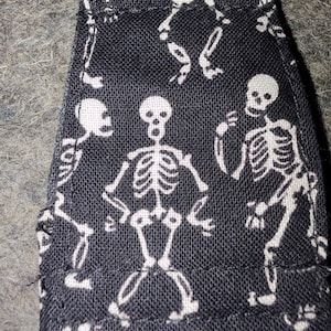 May include: A black fabric strap featuring a repeating white skeleton print. The textile has a visible woven texture and includes a metal ring attachment at the top edge. The skeletons are depicted in various poses across the surface of the material.