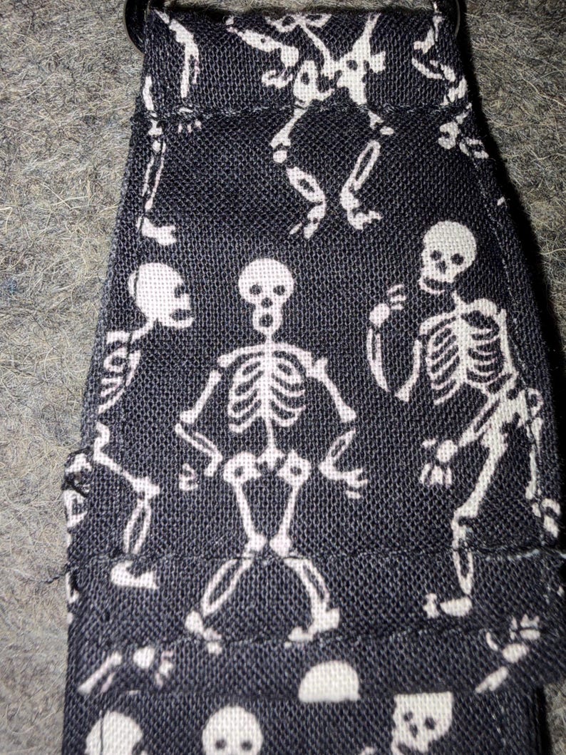 May include: A black fabric strap featuring a repeating white skeleton print. The textile has a visible woven texture and includes a metal ring attachment at the top edge. The skeletons are depicted in various poses across the surface of the material.