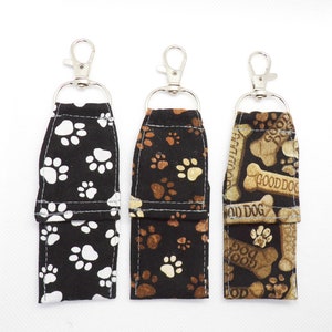 May include: Three keychains with different dog-themed fabric designs. The first keychain has a black background with white paw prints. The second keychain has a brown background with tan paw prints. The third keychain has a brown background with tan bones and the text "Good Dog".