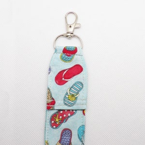 May include: A light blue fabric keychain with a silver clip. The fabric is printed with a pattern of colorful flip flops.