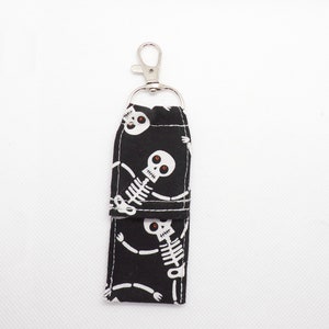 May include: A black fabric lip balm holder featuring a repeating white skeleton pattern with red eyes. The rectangular pouch has a fold-over flap and is attached to a silver-toned metal lobster claw clasp and a circular split ring for attachment.