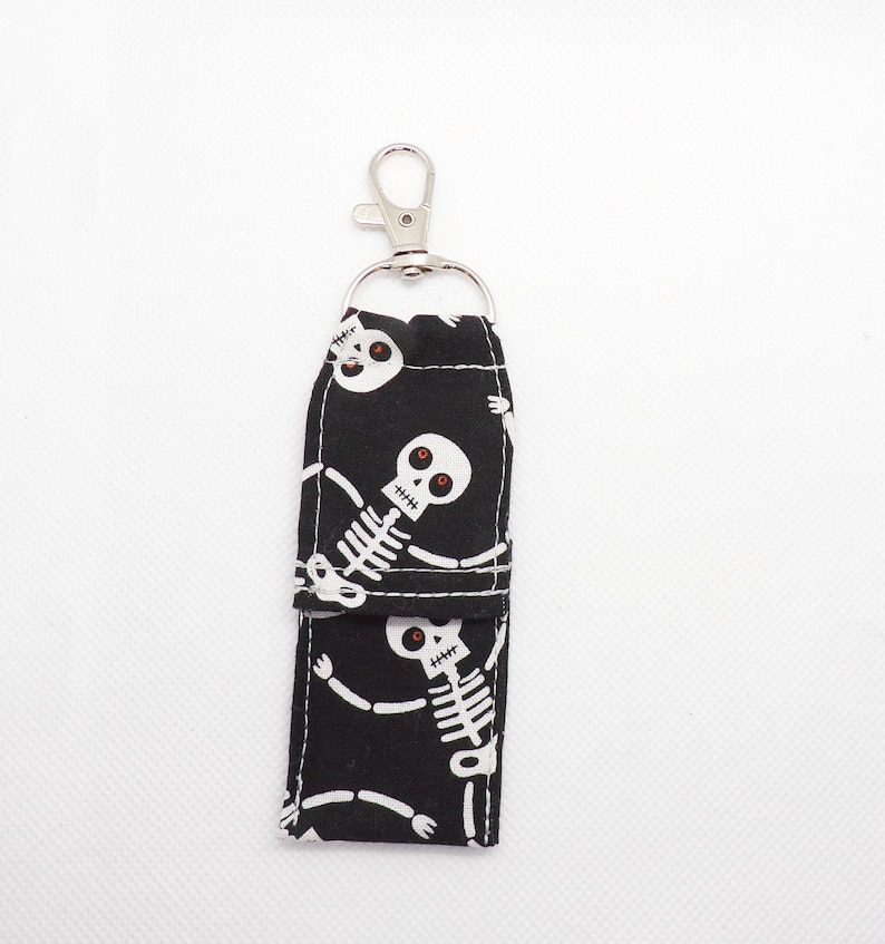 May include: A black fabric lip balm holder featuring a repeating white skeleton pattern with red eyes. The rectangular pouch has a fold-over flap and is attached to a silver-toned metal lobster claw clasp and a circular split ring for attachment.