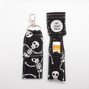 May include: Two black fabric lip balm holders featuring a white skeleton pattern with red eyes. One holder is closed with a silver metal lobster clasp, while the other is open, revealing a lip balm tube and a white circular label reading MY CHAP WRAP.
