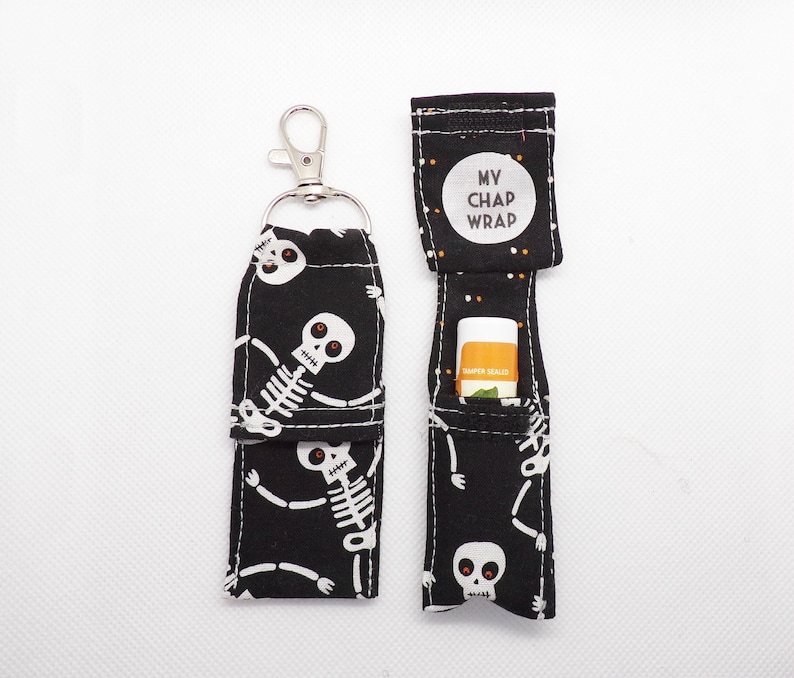 May include: Two black fabric lip balm holders featuring a white skeleton pattern with red eyes. One holder is closed with a silver metal lobster clasp, while the other is open, revealing a lip balm tube and a white circular label reading MY CHAP WRAP.