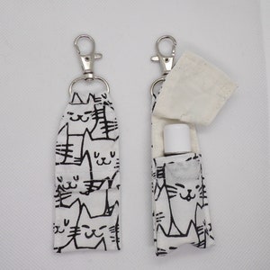 May include: Two white fabric keychain holders with black cat illustrations. Each holder has a silver clip and a small pocket for a lip balm or hand sanitizer.