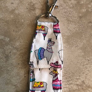 May include: A white fabric keychain with a silver clip. The fabric has a pattern of colorful llamas wearing hats and ponchos.