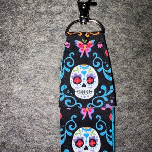 May include: A fabric keychain holder featuring a black background with a colorful pattern of white sugar skulls, pink and blue butterflies, and blue swirling filigree designs. The holder includes a silver-toned metal lobster clasp and ring.