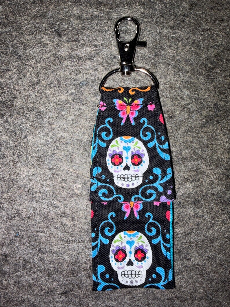 May include: A fabric keychain holder featuring a black background with a colorful pattern of white sugar skulls, pink and blue butterflies, and blue swirling filigree designs. The holder includes a silver-toned metal lobster clasp and ring.
