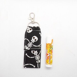 May include: A black fabric lip balm holder featuring a white skeleton pattern with orange eyes, attached to a silver metal lobster clasp. Next to it is a white tube of citrus lip balm with orange slices and the text Citrus with Vitamin E.