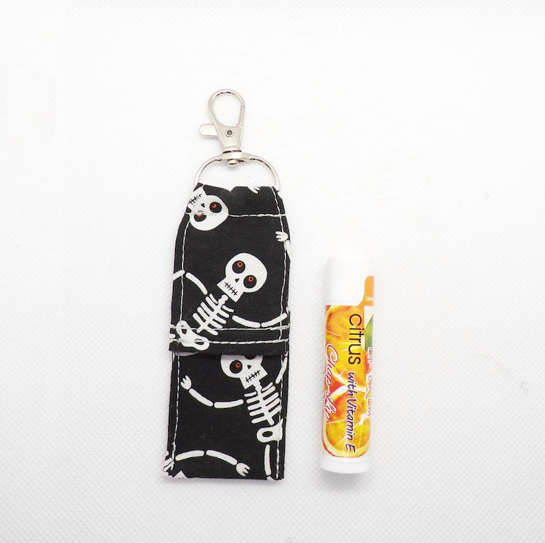 May include: A black fabric lip balm holder featuring a white skeleton pattern with orange eyes, attached to a silver metal lobster clasp. Next to it is a white tube of citrus lip balm with orange slices and the text Citrus with Vitamin E.