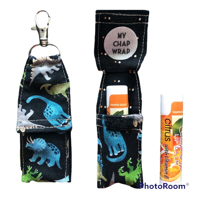K&ouml;nnte beinhalten: Ein schwarzes Stoffetui mit Dinosauriermuster, Rei&szlig;verschluss und silbernem Clip. Das Etui ist f&uuml;r ein Lippenbalsam-R&ouml;hrchen konzipiert. Das Etui ist ge&ouml;ffnet und zeigt ein Lippenbalsam-R&ouml;hrchen mit der Aufschrift "Citrus Lip Balm with Vitamin E". Das Etui hat ein wei&szlig;es Etikett mit der Aufschrift "My Chap Wrap".