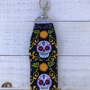 May include: A black and white fabric keychain with a sugar skull design. The skull has purple and red details and is surrounded by yellow and green floral patterns. The keychain has a silver clip.