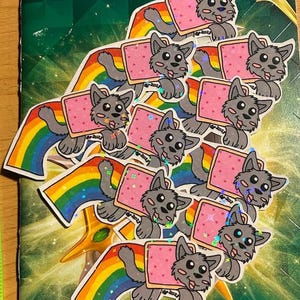 Puede incluir: Un conjunto de nueve pegatinas con un personaje de lobo gris que sostiene una rebanada de pan rosa y blanco con el texto "Nyan Cat" en ella. El lobo está montando un arcoíris con detalles de purpurina. Las pegatinas están sobre un fondo verde y dorado.
