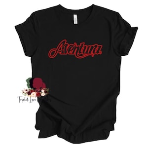 May include: Black t-shirt with the red word "Aventura" in a cursive font. The shirt has short sleeves and a crew neck. A floral design is visible at the bottom left of the shirt.