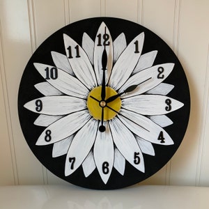 May include: A black and white daisy clock with black numbers and hands. The daisy is painted on a black background.