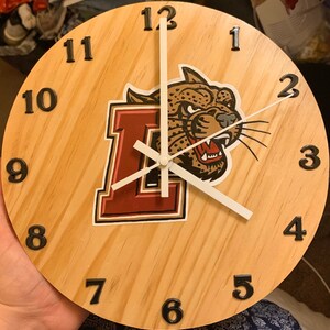 May include: A wooden clock with black numbers 1 through 12 and a white face. The clock has a red and white letter "L" and a brown and black jaguar mascot with a white background. The clock has white hands.