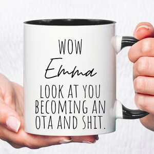May include: White coffee mug with a black rim and handle. The mug has black text that reads "WOW Emma LOOK AT YOU BECOMING AN OTA AND SHIT."
