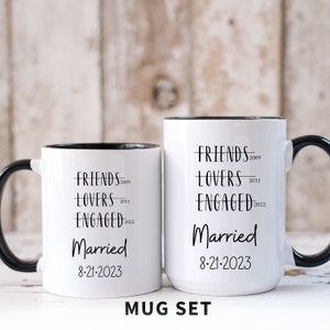 May include: A set of two white mugs with black rims and handles. The mugs have black text that reads "FRIENDS 2009", "LOVERS 2011", "ENGAGED 2022", and "Married 8-21-2023". The mugs are on a white wooden surface.