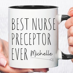 May include: White ceramic mug with a black interior and handle. The mug features the text "BEST NURSE PRECEPTOR EVER Michelle" in black, with a heart symbol. The mug is held by a person.