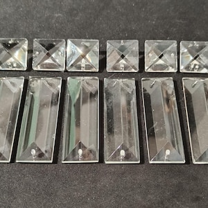 Könnte beinhalten: Eine Sammlung klarer, facettierter Kristall-Kronleuchterprismen. Die Anordnung umfasst quadratische und rechteckige Formen, die Licht reflektieren. Die Prismen sind für dekorative Beleuchtung konzipiert und auf einer dunklen Oberfläche angeordnet.