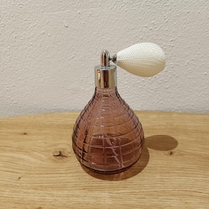 Könnte beinhalten: Eine violette Glasflasche mit strukturierter Oberfläche und einem weißen Plastikzerstäuber. Die Flasche steht auf einer Holzoberfläche.