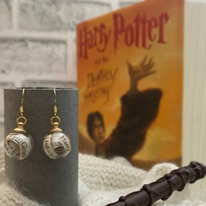 May include: A pair of gold dangle earrings with glass orbs containing small pieces of paper with the words "Harry Potter", "Ginny", "Hedwig", and "Time" printed on them. The earrings are displayed on a gray stand in front of a Harry Potter book.