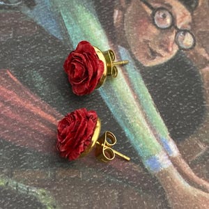 May include: A pair of red rose stud earrings with gold-colored posts and settings. The earrings are set against a colorful background with a blurred illustration.