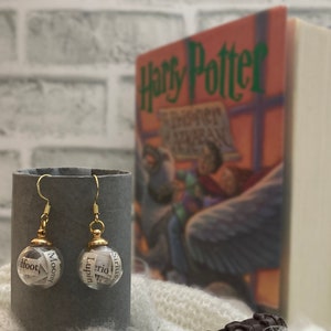 May include: A pair of gold-toned earrings with small glass globes filled with rolled up paper. The paper has words printed on it, including "Sirius", "Lupin", "Moony", and "Dfoot". The earrings are in front of a copy of the book "Harry Potter and the Prisoner of Azkaban".
