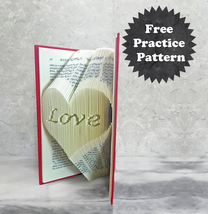 May include: A red book with a white heart shaped book fold inside. The heart has the word "Love" written inside. The book is on a gray surface. Free practice pattern.