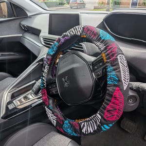 May include: A black steering wheel cover featuring a repeating pattern of stylized leaves in vibrant shades of pink, bright blue, and white. The fabric cover is elasticized and fitted snugly around the circular steering wheel of a vehicle.