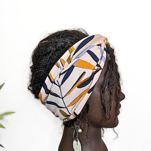 May include: A beige headband featuring a leaf design in navy, yellow, and green. The headband is worn on a dark-skinned mannequin head. A dangling earring with a leaf charm is also visible.