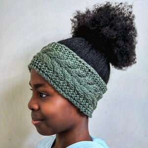 Puede incluir: Perfil de una persona que lleva una diadema verde de ganchillo con un diseño de punto de cable. La diadema es de color verde apagado y cubre la frente y las orejas. El pelo de la persona es negro y está recogido en un moño.