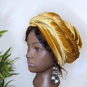 May include: A gold velvet turban headwrap. The turban is tied in a knot on top of the head and drapes down the sides.