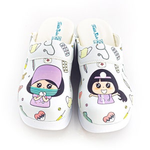 Puede incluir: Un par de zuecos blancos con un diseño de médico de dibujos animados. Los zuecos presentan un patrón azul claro y blanco con un estetoscopio, píldoras y otros símbolos médicos. Los zuecos tienen una suela blanca y una correa blanca.