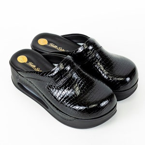 Peut inclure: Paire de sabots en cuir noir à motif peau de serpent. Les sabots ont une épaisse semelle plateforme et un logo doré qui indique "Textile Shoes".