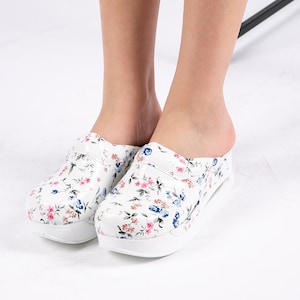 May include: White clogs with a floral print featuring pink, blue, and brown flowers. The clogs have a thick white sole.