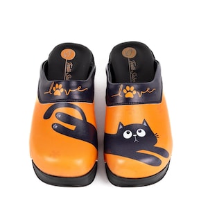 May include: A pair of orange clogs with a black cat design. The clogs have a black sole and a black top with the word "love" and a paw print. The brand name "Terlik Sabo" is printed on the top.