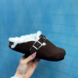 Pode incluir: Sapatilha de couro camurça marrom com forro de pele sintética branca e fivela prateada. A sapatilha está sendo segurada por uma mão contra um fundo azul.