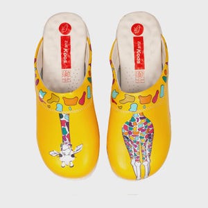Pode incluir: Tamancos amarelos com um design de girafa. Os tamancos têm um interior branco com o texto "Zue Koos" impresso na lateral. O design da girafa apresenta um padrão colorido no corpo e no pescoço da girafa.