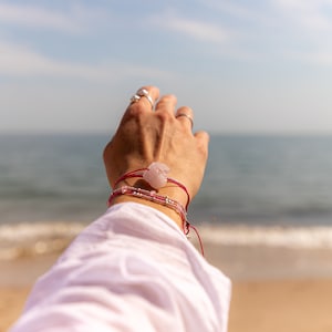 Op de afbeelding: Een hand met een rood koordarmband met een roze stenen bedel en een zilveren ring reikt naar een wazige achtergrond van een strand en de oceaan.