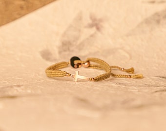 May include: A gold cross charm bracelet with a beige braided cord and gold beads. The bracelet has a tassel on one end.