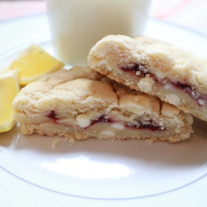 Pode incluir: Close-up de um prato branco com duas metades de uma barra de biscoito, mostrando um recheio vermelho e gotas de chocolate branco. Fatias de limão e um copo de leite estão no fundo. A barra de biscoito tem uma textura macia e esfarelada.