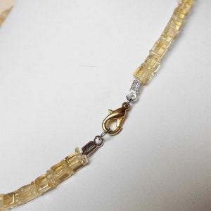 May include: A gold necklace with a clasp made of silver metal. The necklace is made of small, square-shaped, light brown stones.